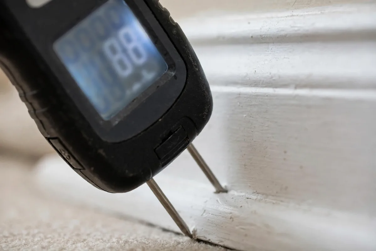Pin-type moisture meter pressed against a baseboard showing an elevated moisture reading